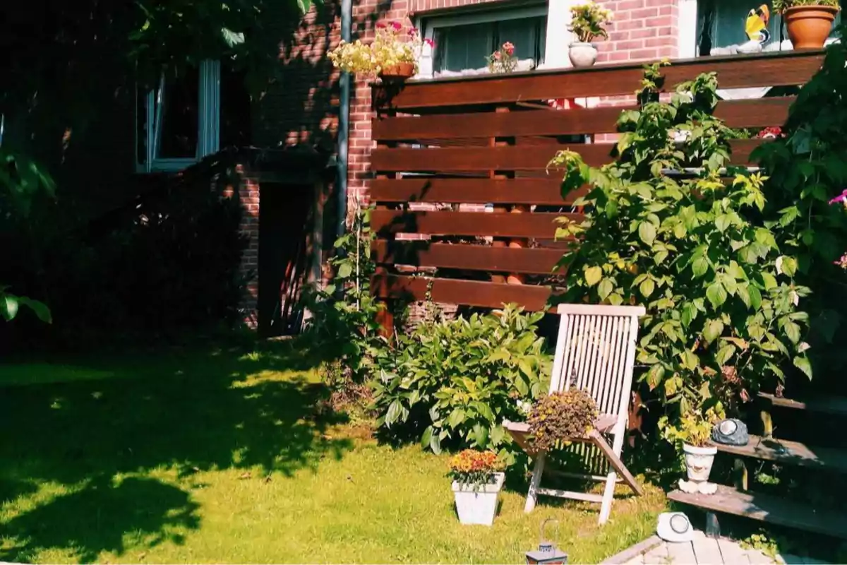 Silla de madera en un jardín soleado junto a plantas y macetas frente a una cerca de madera y una casa de ladrillo