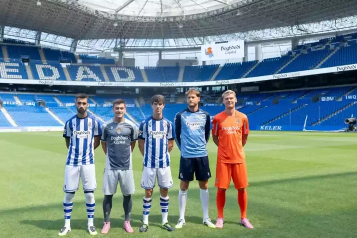 Cinco futbolistas posan en el campo de un estadio vacío con diferentes uniformes del equipo Real Sociedad