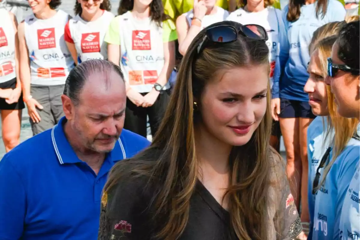 Joven con gafas de sol en la cabeza y cabello largo sonríe mientras camina entre un grupo de personas vestidas con camisetas deportivas Joven con gafas de sol en la cabeza y cabello largo sonríe mientras camina entre un grupo de personas vestidas con camisetas deportivas