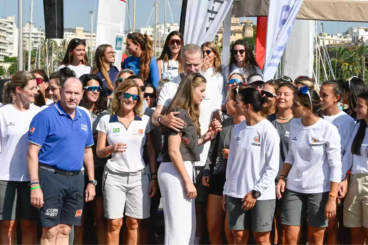 Un grupo numeroso de personas vestidas con ropa deportiva se reúne al aire libre en un puerto deportivo con veleros y edificios al fondo, algunas personas sonríen y otras conversan entre sí. Un grupo numeroso de personas vestidas con ropa deportiva se reúne al aire libre en un puerto deportivo con veleros y edificios al fondo, algunas personas sonríen y otras conversan entre sí.