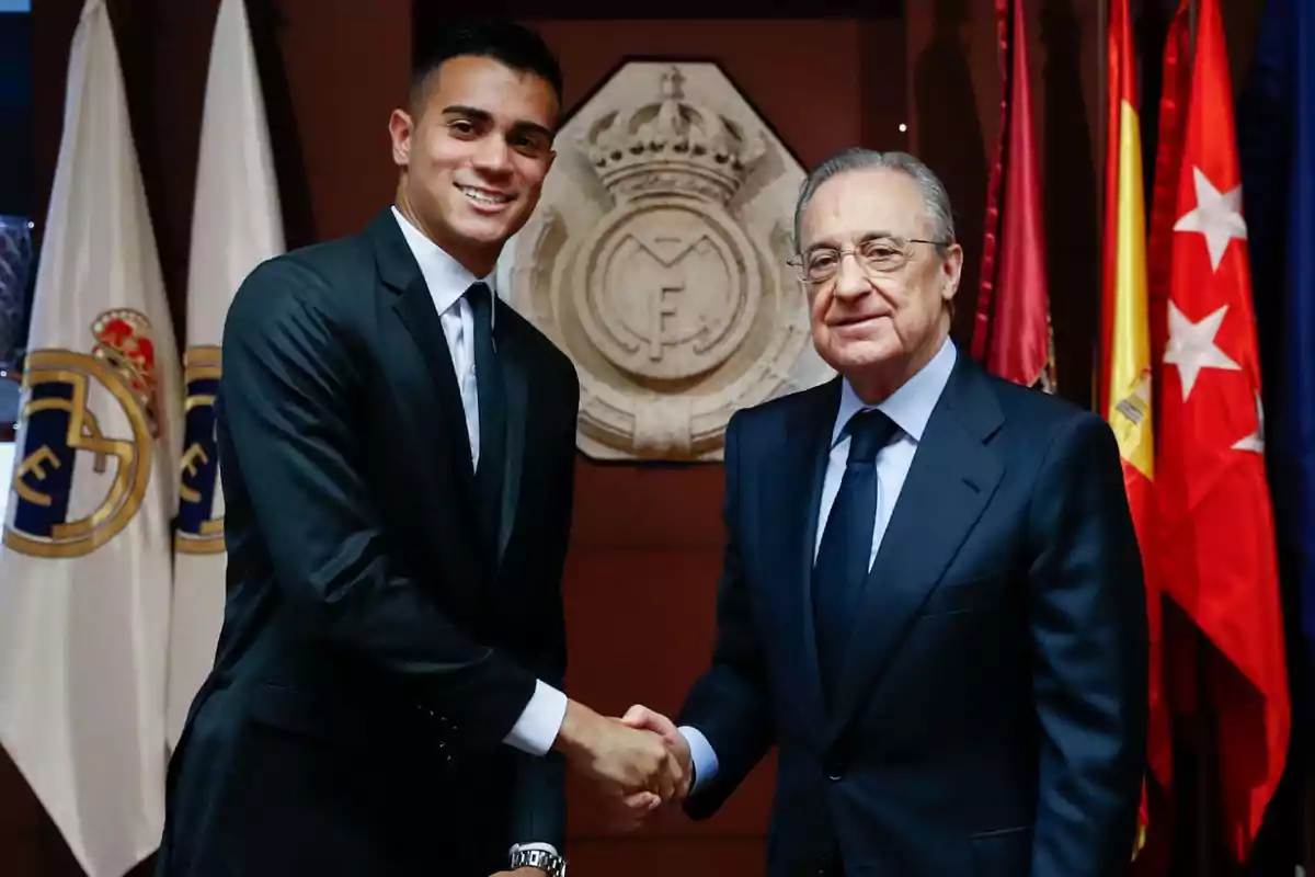 Dos hombres con traje se dan la mano frente a banderas y el escudo del Real Madrid.