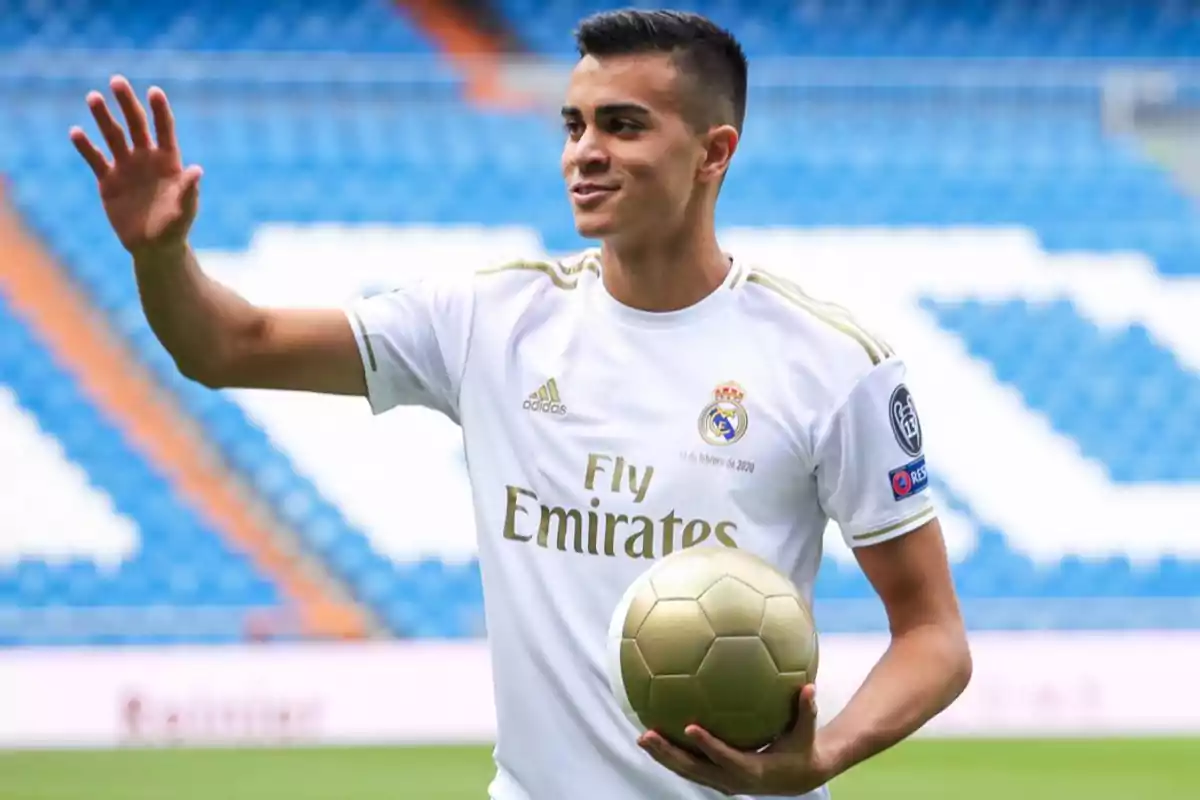 Joven futbolista con uniforme blanco del Real Madrid sostiene un balón dorado y saluda en un estadio vacío Joven futbolista con uniforme blanco del Real Madrid sostiene un balón dorado y saluda en un estadio vacío