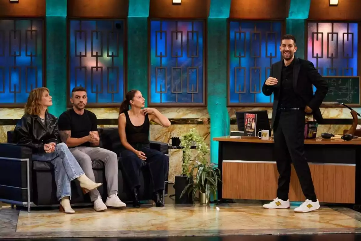 Tres personas sentadas en un sofá conversan mientras un hombre de pie sonríe junto a un escritorio en un set de televisión con luces azules y verdes de fondo Tres personas sentadas en un sofá conversan mientras un hombre de pie sonríe junto a un escritorio en un set de televisión con luces azules y verdes de fondo