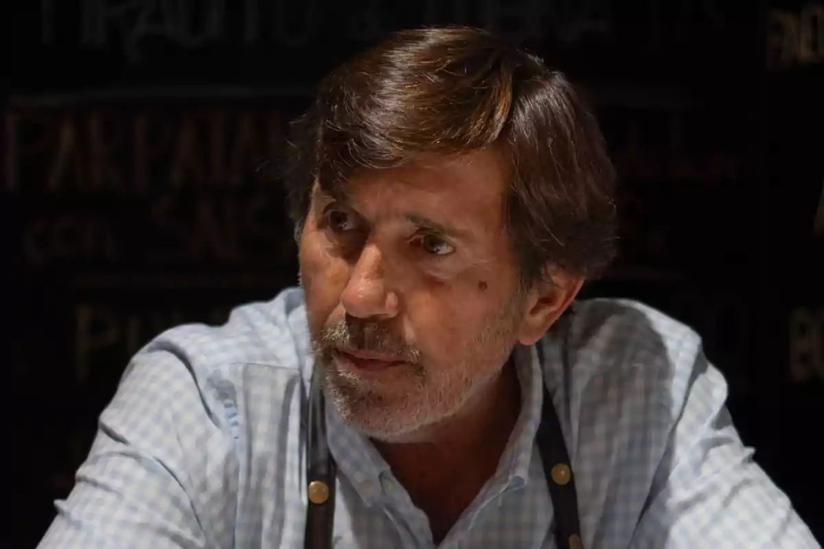 Hombre de mediana edad con barba y cabello castaño, usando una camisa de cuadros y tirantes, sentado en un ambiente interior con fondo oscuro. Hombre de mediana edad con barba y cabello castaño, usando una camisa de cuadros y tirantes, sentado en un ambiente interior con fondo oscuro.