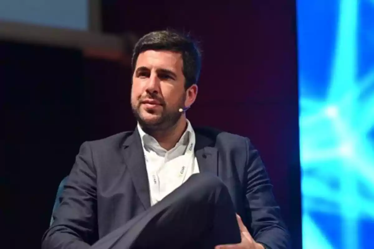 Hombre con barba y cabello oscuro usando traje y micrófono de diadema sentado en un escenario con fondo azul iluminado Hombre con barba y cabello oscuro usando traje y micrófono de diadema sentado en un escenario con fondo azul iluminado