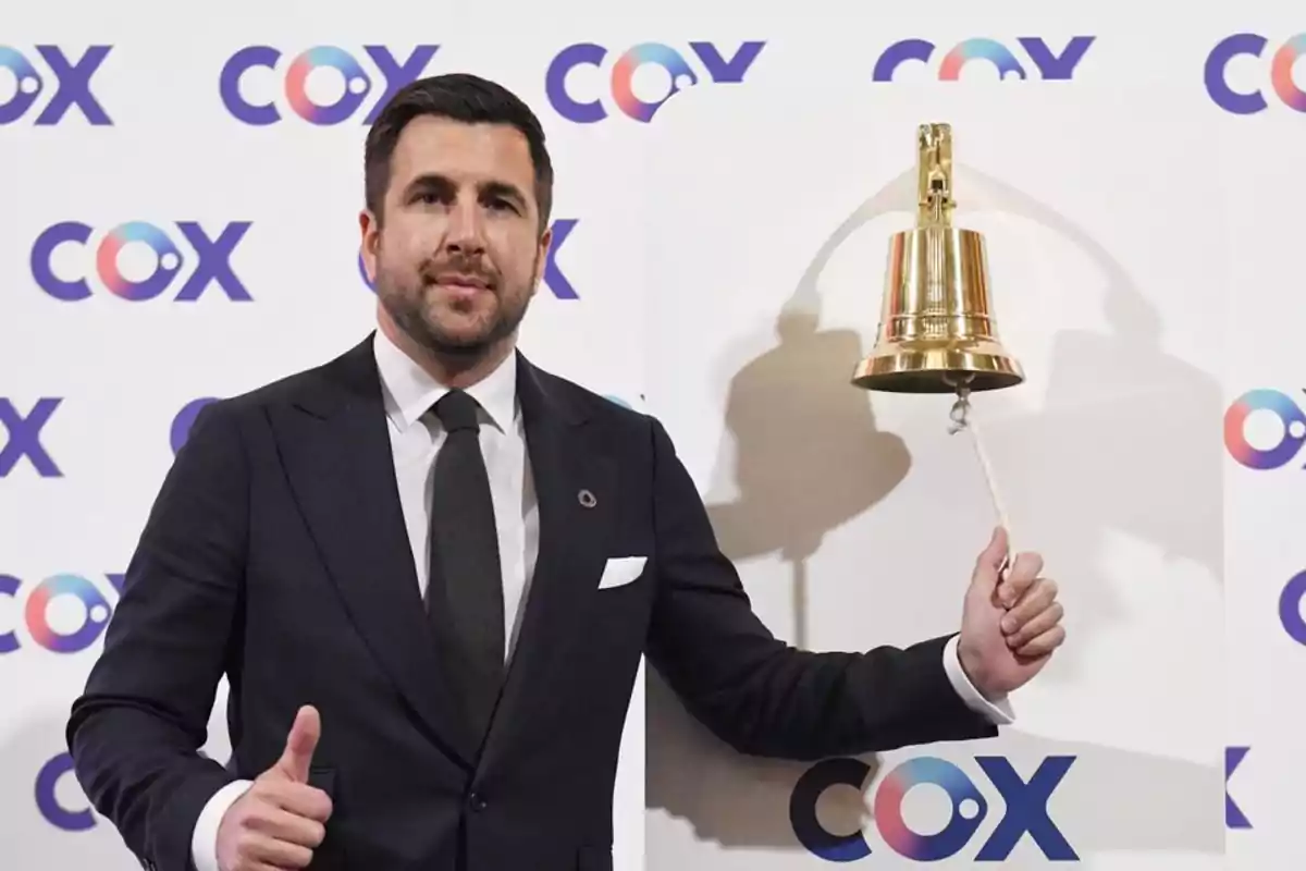 Hombre de traje tocando una campana dorada mientras sonríe y hace un gesto de aprobación con el pulgar frente a un fondo con el logo de COX
