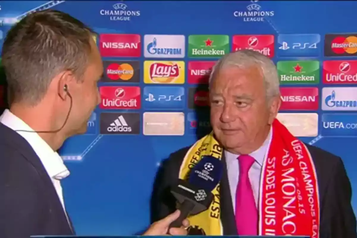 Dos hombres conversan durante una entrevista en un evento de la Champions League uno de ellos lleva una bufanda con los colores de dos equipos y sostiene un micrófono con el logo del torneo mientras de fondo se ven logotipos de patrocinadores Dos hombres conversan durante una entrevista en un evento de la Champions League uno de ellos lleva una bufanda con los colores de dos equipos y sostiene un micrófono con el logo del torneo mientras de fondo se ven logotipos de patrocinadores
