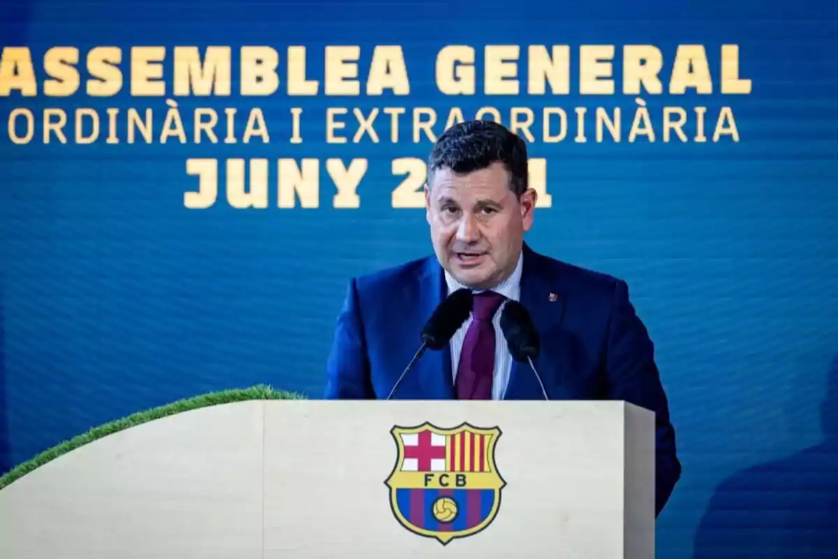 Hombre con traje hablando en un podio con el escudo del FC Barcelona y un fondo azul con letras amarillas que anuncian una asamblea general en junio de 2021 Hombre con traje hablando en un podio con el escudo del FC Barcelona y un fondo azul con letras amarillas que anuncian una asamblea general en junio de 2021