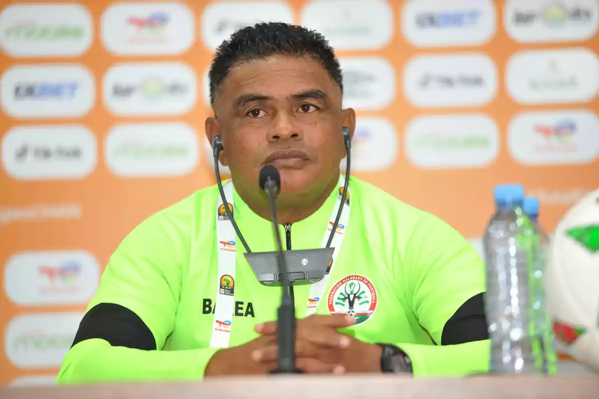 Entrenador de fútbol con uniforme verde fosforescente sentado en una conferencia de prensa frente a un micrófono con fondo de logotipos y una botella de agua a un lado