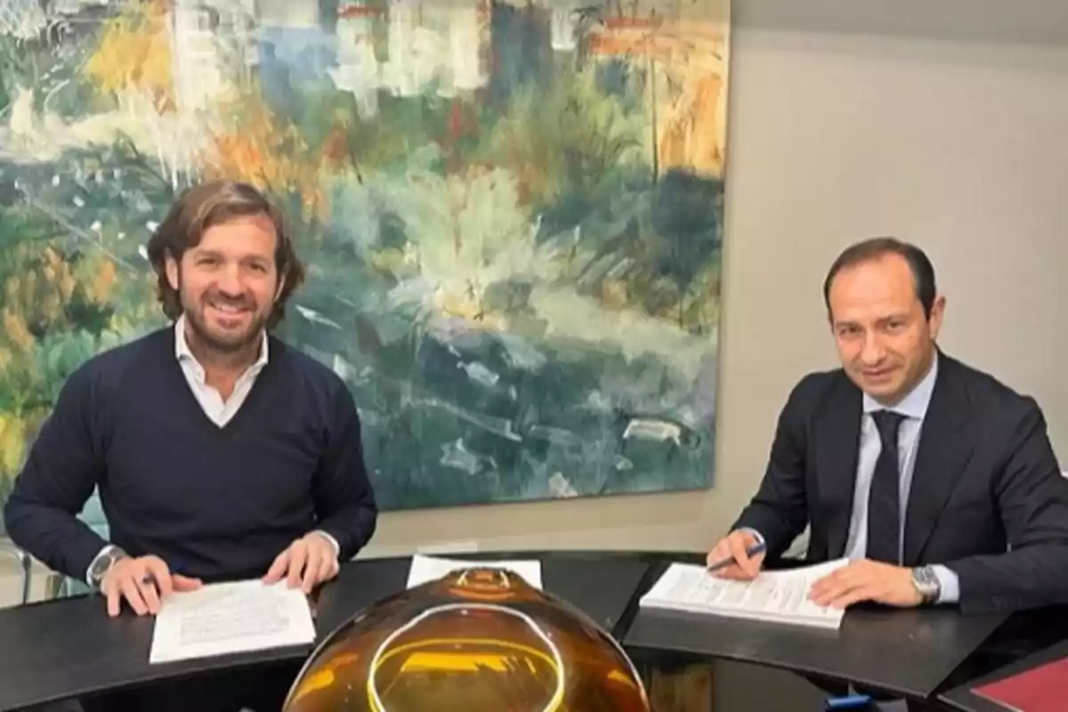 Dos hombres sentados en una mesa firmando documentos, ambos sonríen y llevan ropa formal, al fondo hay una pintura abstracta colorida.