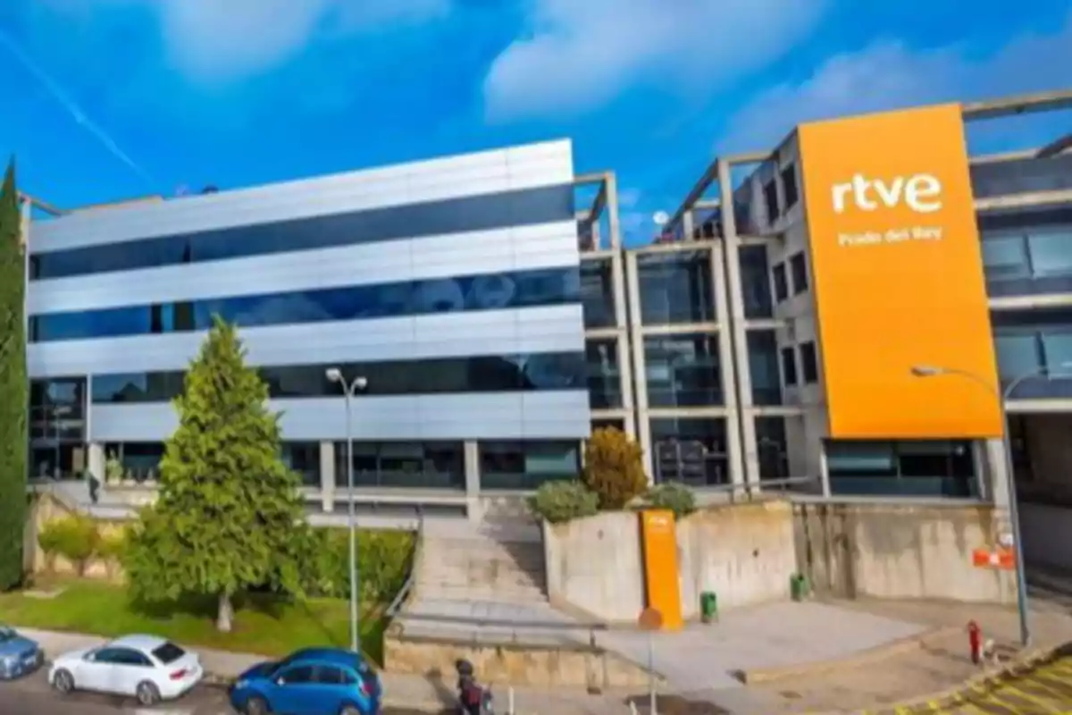 Edificio moderno de RTVE Prado del Rey con fachada de vidrio y paneles metálicos, un gran letrero naranja y varios autos estacionados al frente Edificio moderno de RTVE Prado del Rey con fachada de vidrio y paneles metálicos, un gran letrero naranja y varios autos estacionados al frente