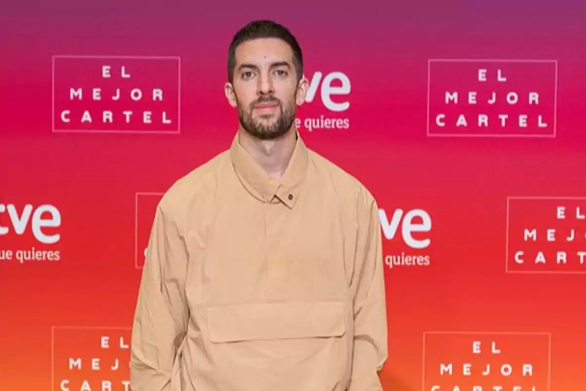 Hombre con barba y cabello corto viste una chaqueta beige posando frente a un fondo rojo con logotipos de RTVE y el texto El Mejor Cartel