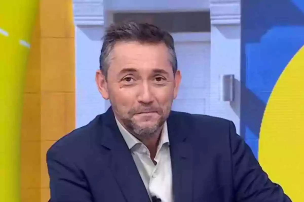 Hombre de cabello corto y barba con saco azul y camisa clara en un estudio de televisión con fondo colorido Hombre de cabello corto y barba con saco azul y camisa clara en un estudio de televisión con fondo colorido