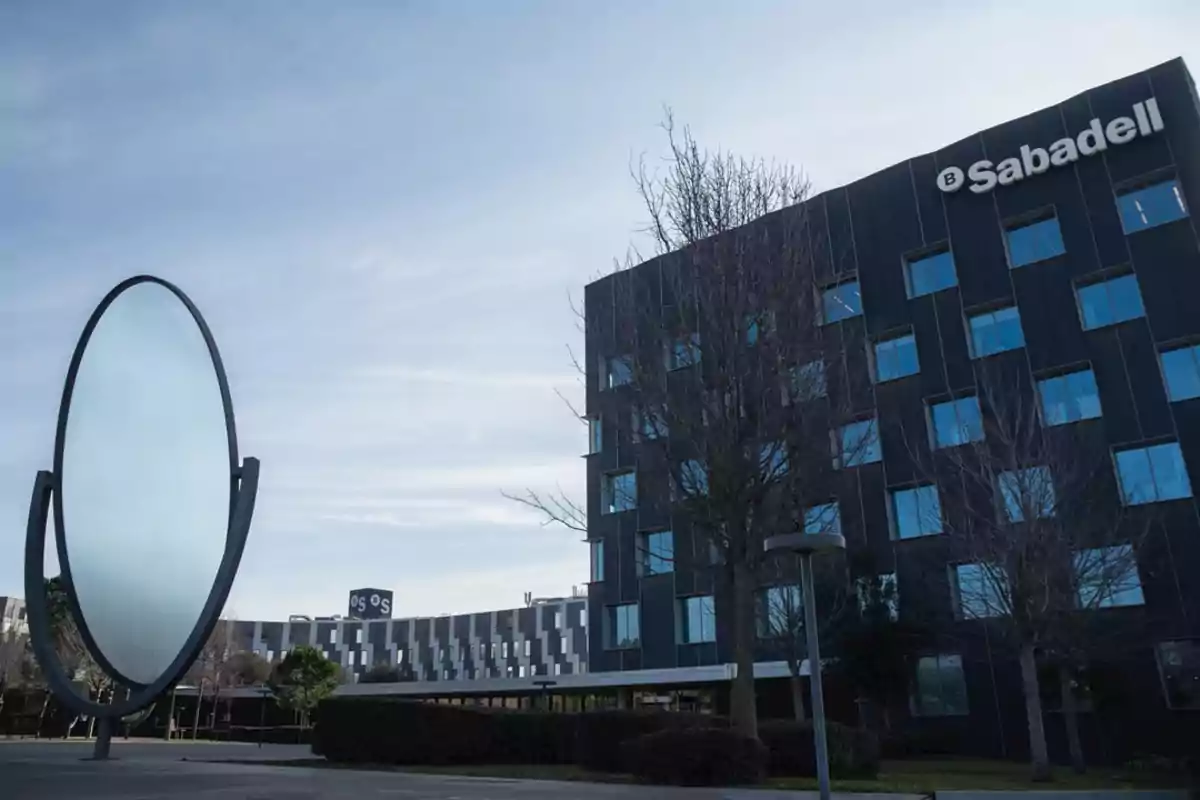Edificio moderno de Sabadell con ventanas azules y una gran escultura circular en el exterior rodeado de árboles sin hojas Edificio moderno de Sabadell con ventanas azules y una gran escultura circular en el exterior rodeado de árboles sin hojas