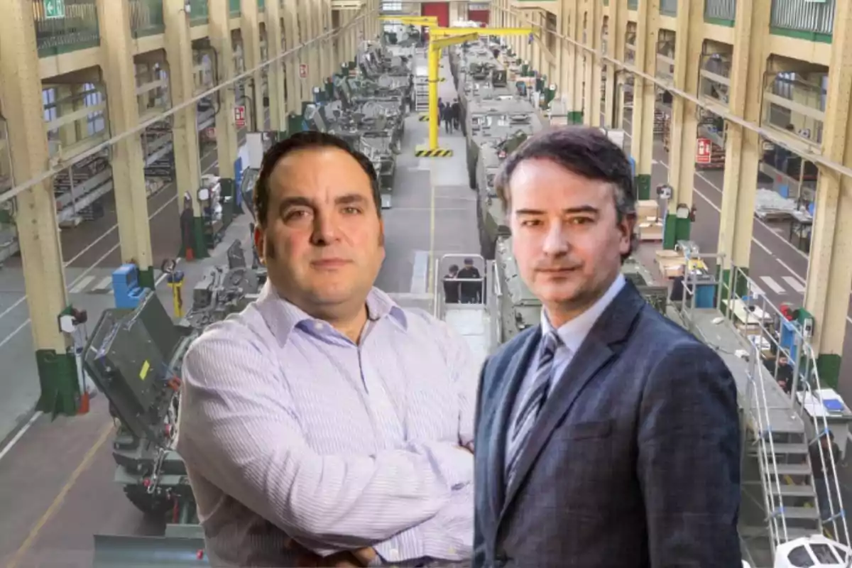 Dos hombres posan al frente de una fábrica industrial con vehículos militares en proceso de ensamblaje.