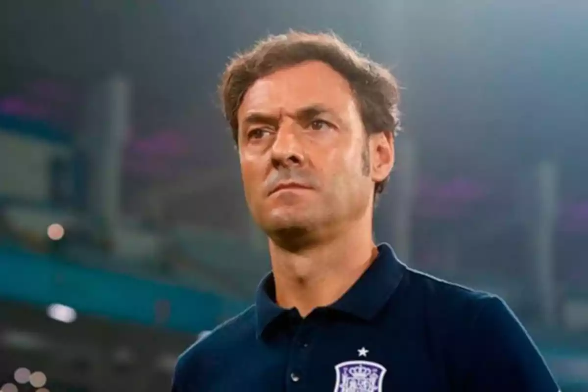 Hombre con expresión seria vistiendo una camiseta azul marino con un escudo en el pecho en un entorno deportivo. Hombre con expresión seria vistiendo una camiseta azul marino con un escudo en el pecho en un entorno deportivo.
