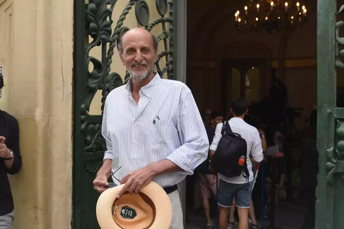 Hombre mayor sonriente con camisa de rayas y sombrero en la mano de pie en la entrada de un edificio con puertas verdes y personas al fondo