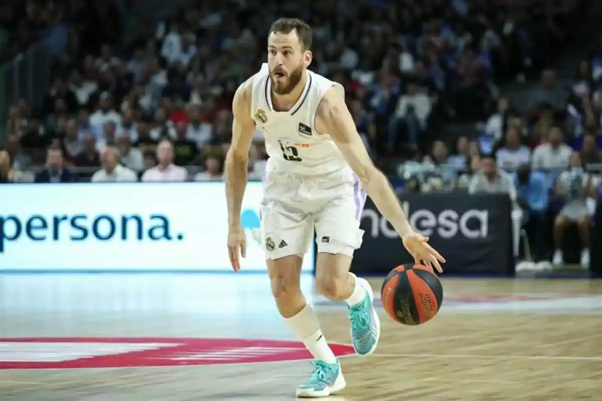 Jugador de baloncesto con uniforme blanco driblando el balón en una cancha durante un partido con público de fondo Jugador de baloncesto con uniforme blanco driblando el balón en una cancha durante un partido con público de fondo