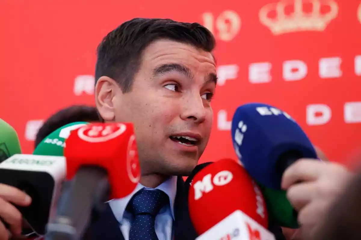 Hombre con traje hablando ante varios micrófonos de diferentes medios de comunicación con fondo rojo