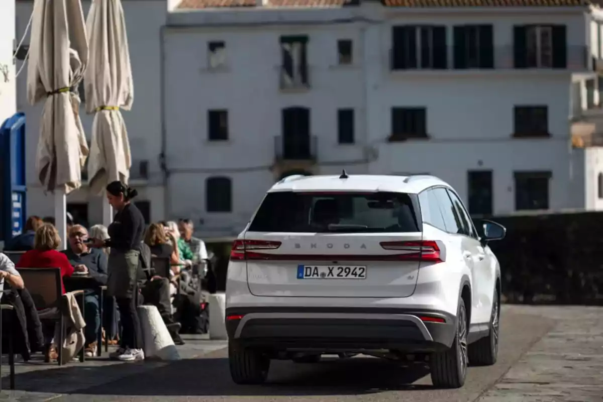 Un coche blanco estacionado cerca de una terraza donde varias personas están sentadas y una camarera toma una orden en un entorno urbano con edificios blancos al fondo Un coche blanco estacionado cerca de una terraza donde varias personas están sentadas y una camarera toma una orden en un entorno urbano con edificios blancos al fondo