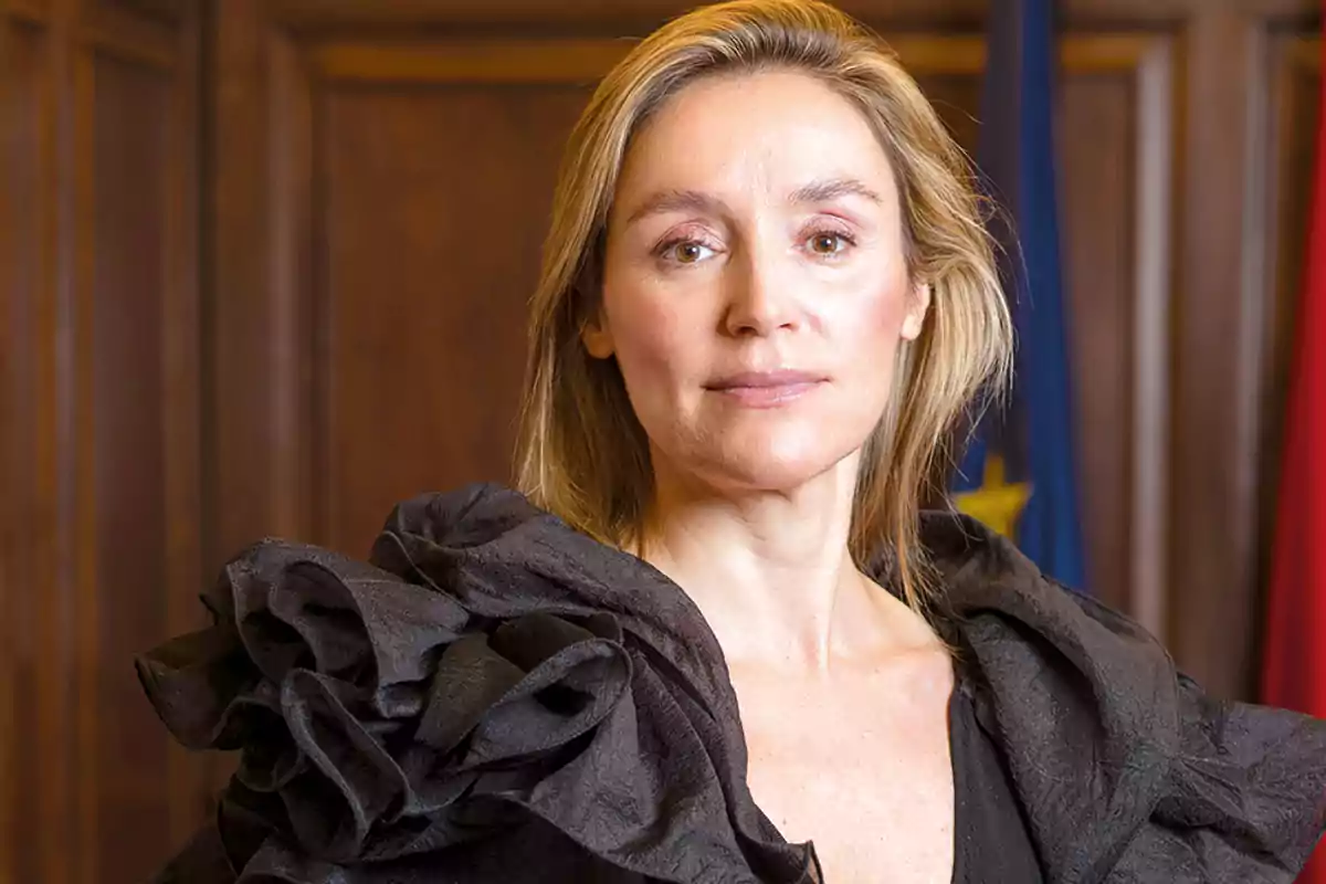Mujer rubia con expresión seria vestida con un atuendo negro elegante y fondo de madera con banderas desenfocadas