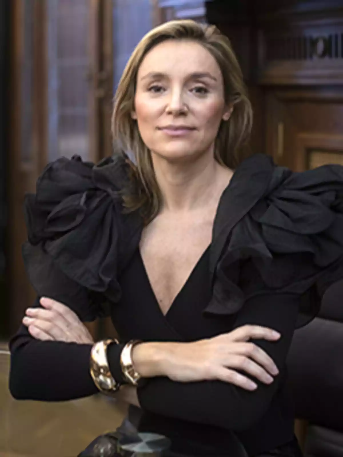 Mujer sentada con los brazos cruzados, vestida de negro con mangas voluminosas y pulseras doradas, en un entorno elegante de madera