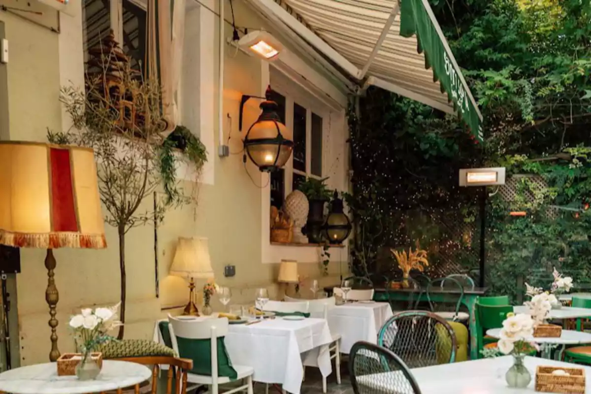 Terraza de restaurante decorada con lámparas vintage, mesas con manteles blancos y flores, rodeada de plantas y luces cálidas Terraza de restaurante decorada con lámparas vintage, mesas con manteles blancos y flores, rodeada de plantas y luces cálidas