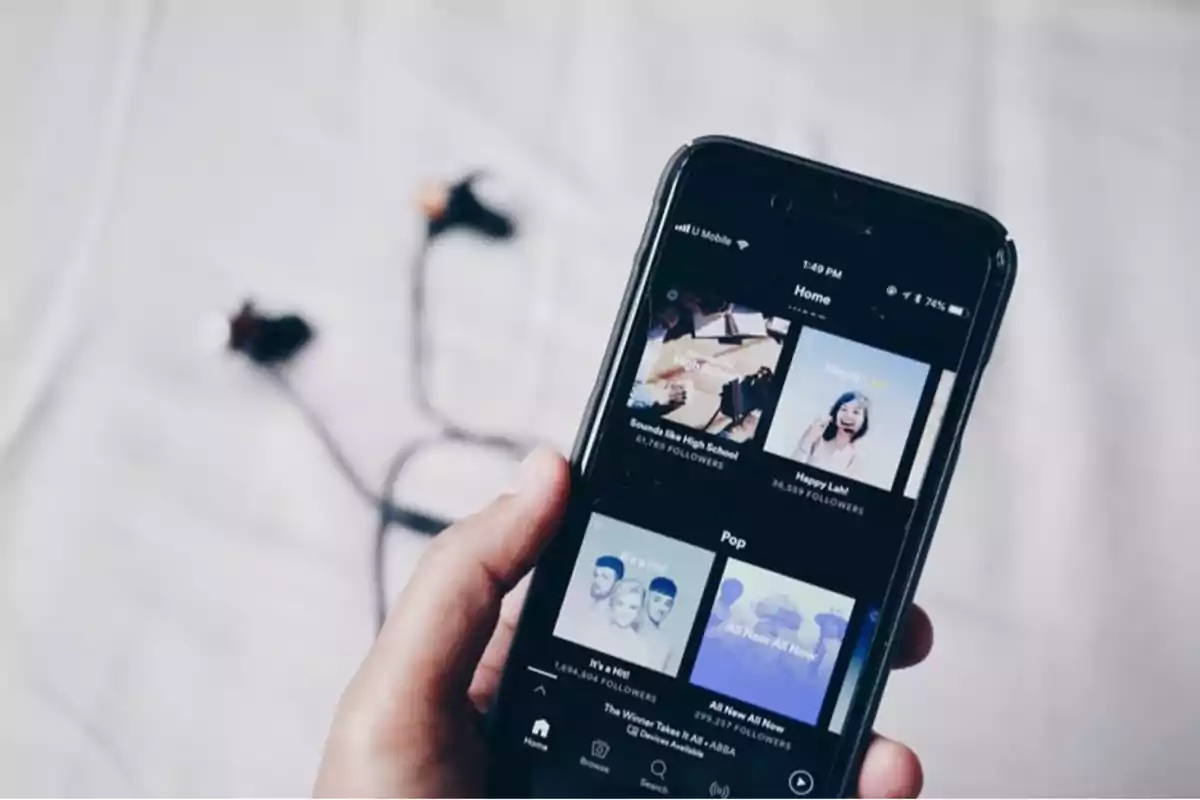 Persona sosteniendo un teléfono móvil con la pantalla mostrando una aplicación de música y unos audífonos desenrollados sobre una superficie blanca