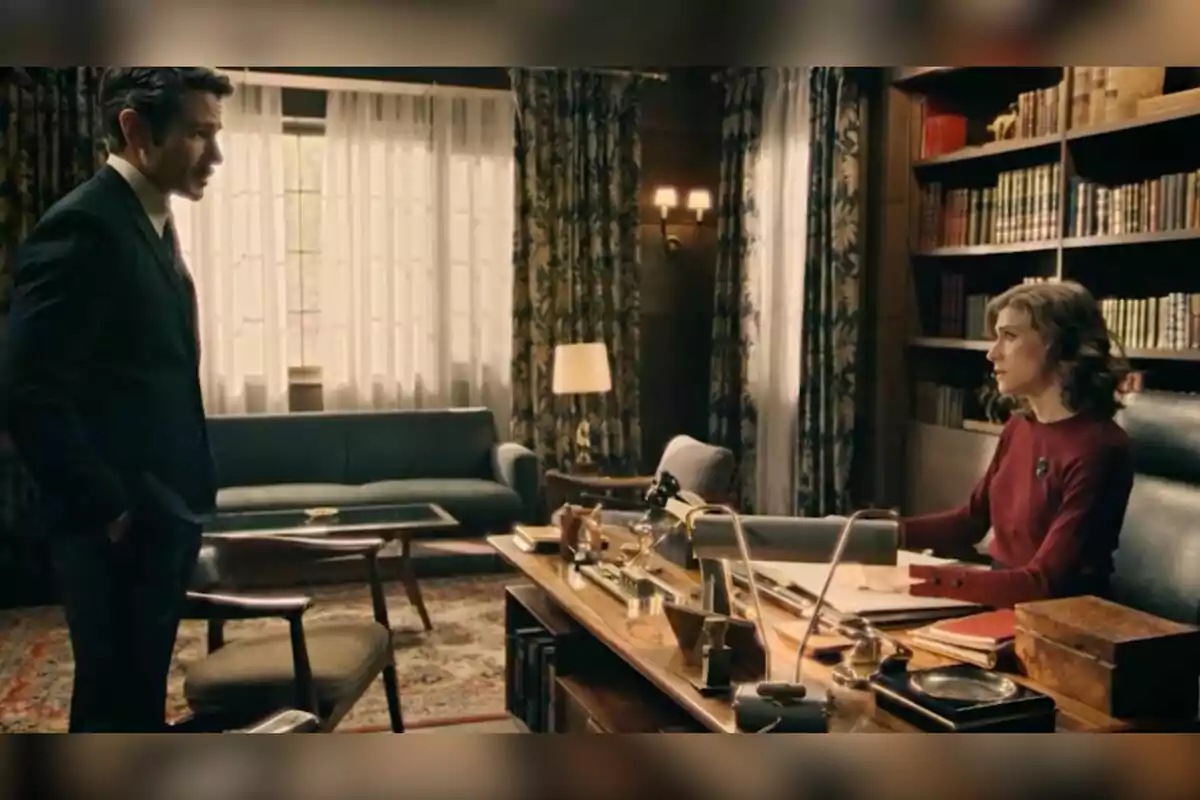 Pelayo de pie y Marta sentada conversan en una oficina elegante con estanterías de libros, cortinas y muebles clásicos. Captura de 'Sueños de libertad' Pelayo de pie y Marta sentada conversan en una oficina elegante con estanterías de libros, cortinas y muebles clásicos. Captura de 'Sueños de libertad'