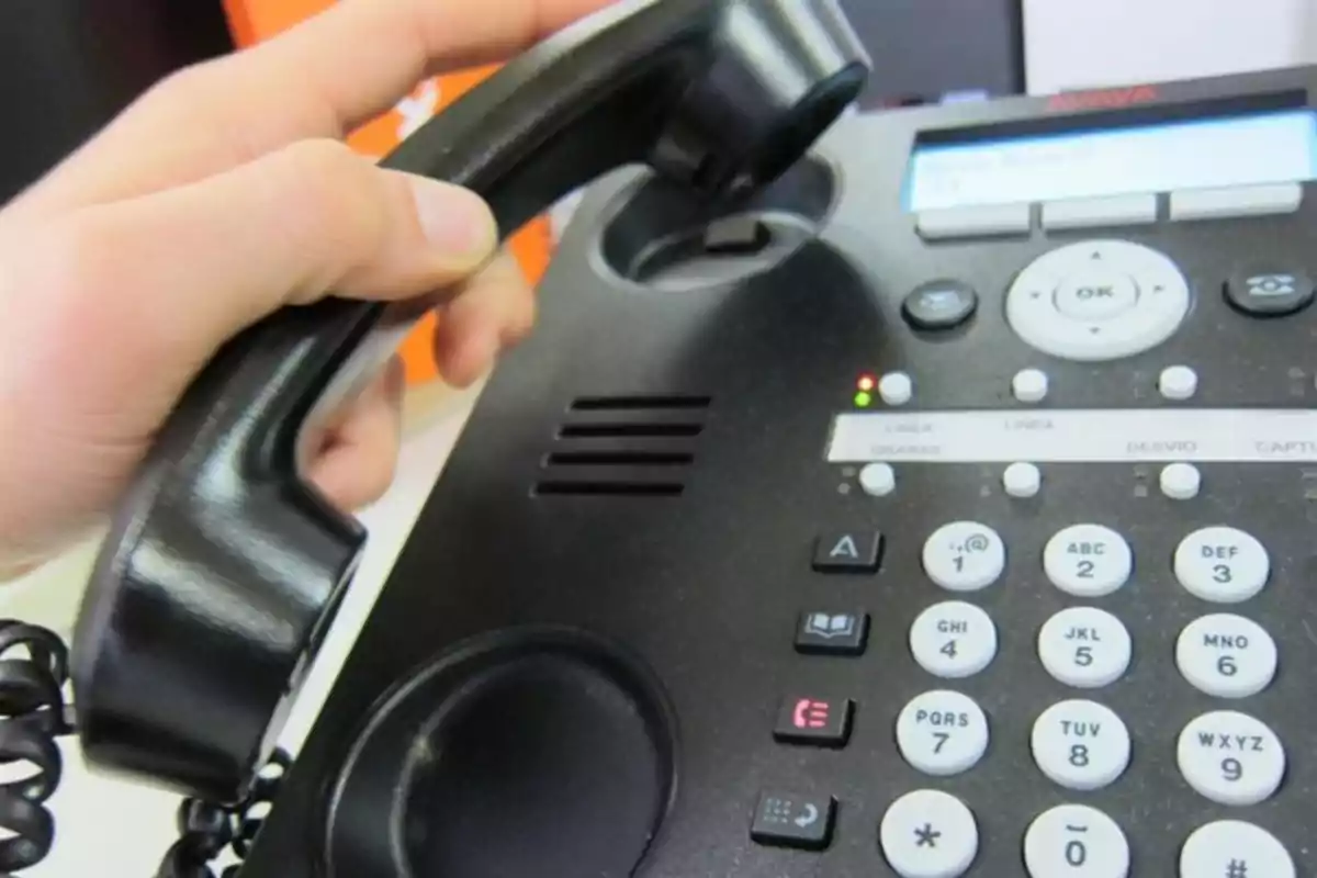 Persona levantando el auricular de un teléfono de oficina negro con botones y pantalla digital