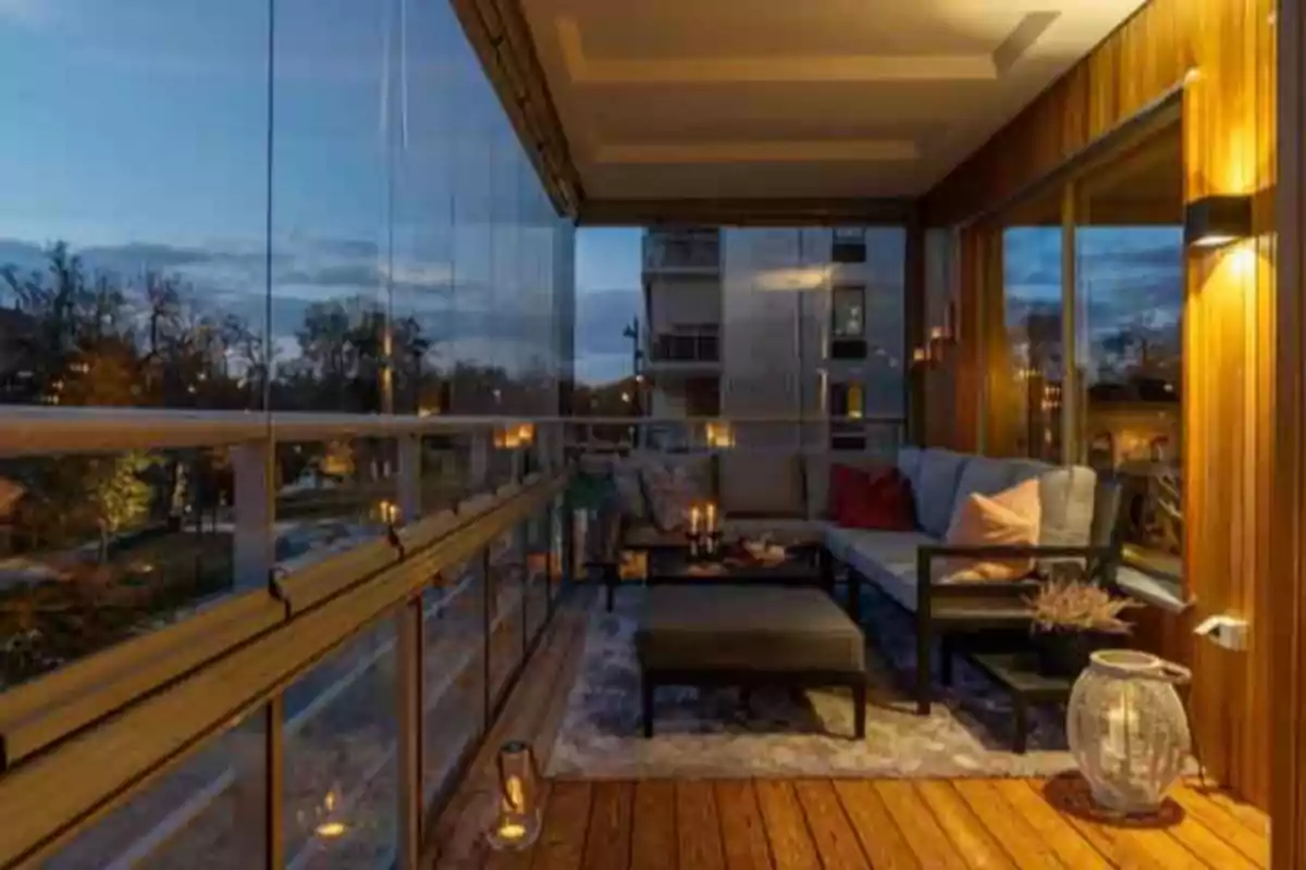 Terraza acristalada al atardecer con sofá en L, cojines, mesa baja, alfombra y luces cálidas creando un ambiente acogedor