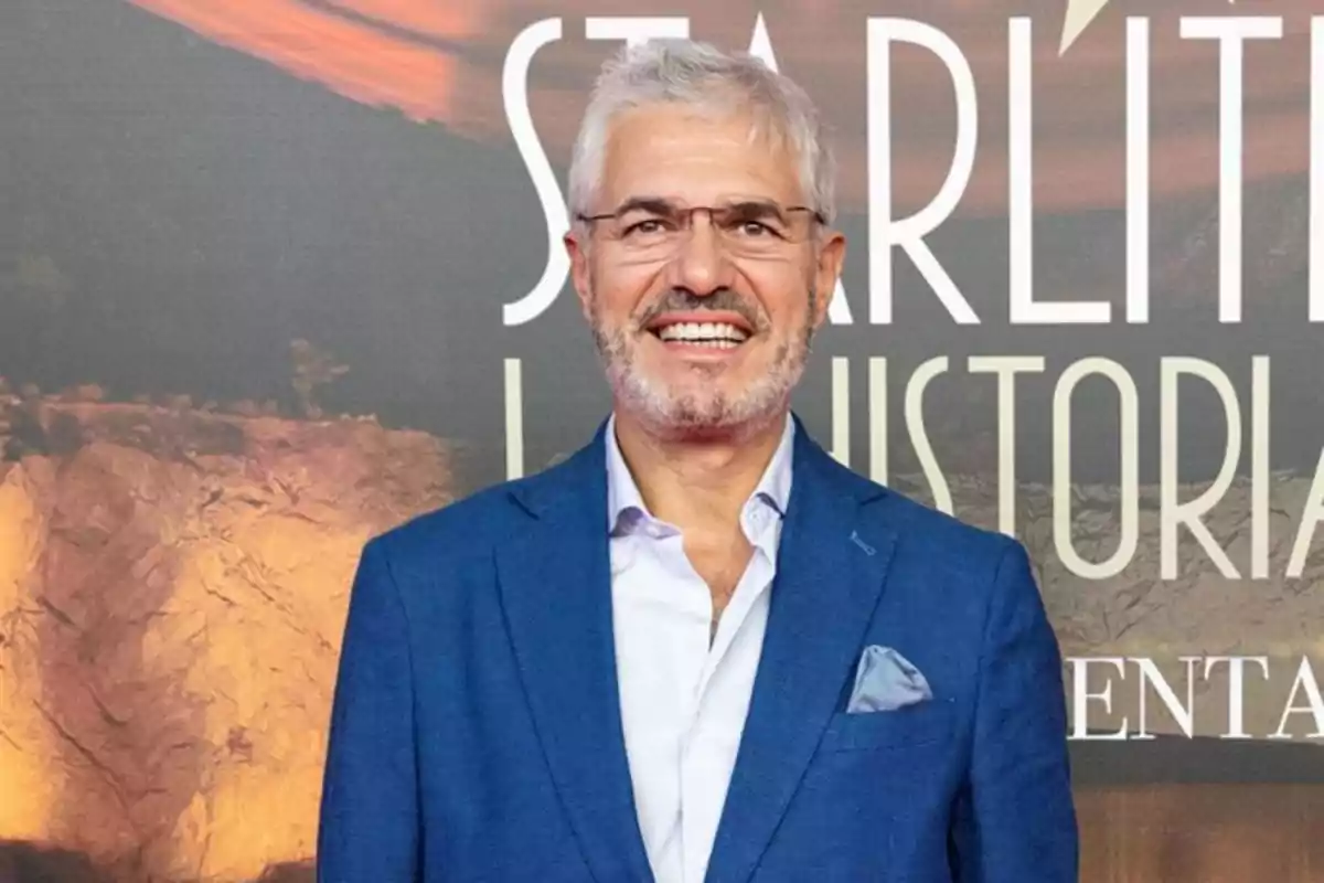 Hombre de cabello canoso y barba corta, sonriente, con gafas y vestido con saco azul y camisa clara, posando frente a un fondo con letras grandes.