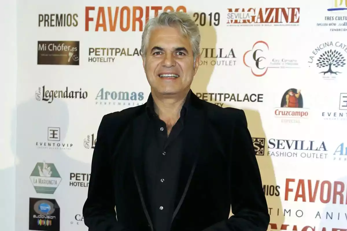 Hombre de cabello canoso y sonrisa vistiendo un traje negro posando frente a un fondo con logotipos de patrocinadores en un evento de premios Hombre de cabello canoso y sonrisa vistiendo un traje negro posando frente a un fondo con logotipos de patrocinadores en un evento de premios