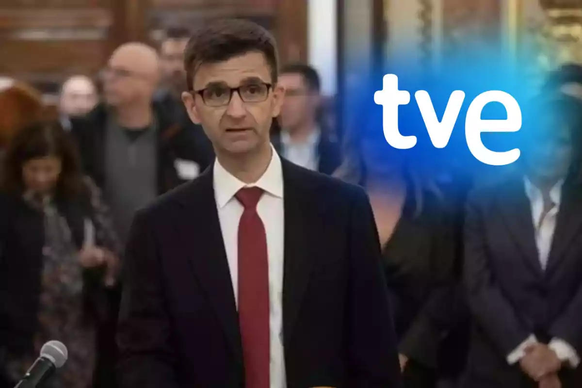 Hombre con gafas y corbata roja hablando frente a un micrófono con el logo de tve sobre un fondo de personas desenfocadas