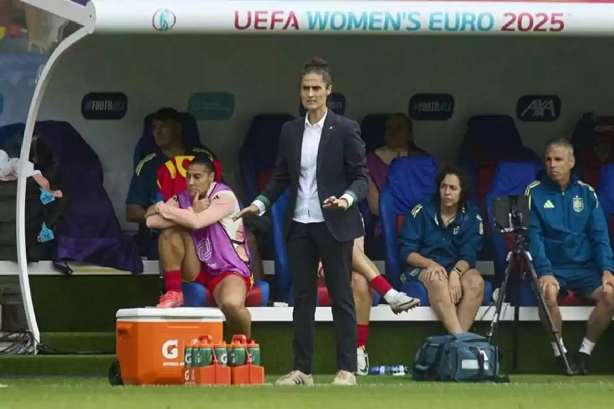 Entrenadora de fútbol de pie frente al banquillo durante un partido del Campeonato Europeo Femenino de la UEFA 2025 con jugadoras y cuerpo técnico sentados detrás de ella Entrenadora de fútbol de pie frente al banquillo durante un partido del Campeonato Europeo Femenino de la UEFA 2025 con jugadoras y cuerpo técnico sentados detrás de ella