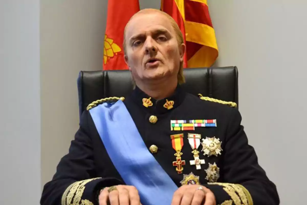 Hombre vestido con uniforme militar con medallas y banda azul sentado frente a dos banderas Hombre vestido con uniforme militar con medallas y banda azul sentado frente a dos banderas