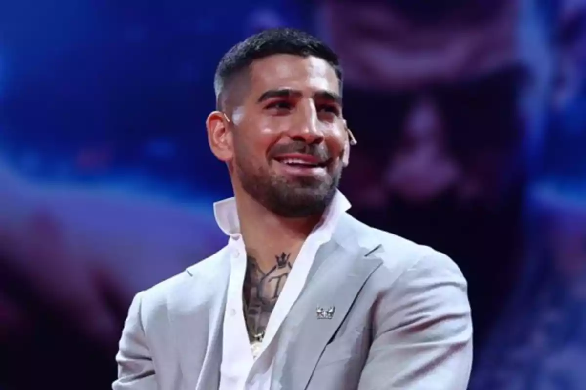 Hombre con barba y cabello corto vestido con saco gris claro y camisa blanca sonriendo en un ambiente iluminado con fondo desenfocado azul y púrpura Hombre con barba y cabello corto vestido con saco gris claro y camisa blanca sonriendo en un ambiente iluminado con fondo desenfocado azul y púrpura