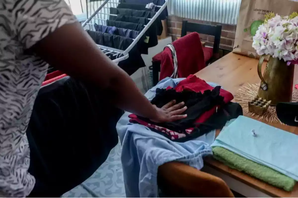 Persona doblando ropa sobre una mesa de madera junto a un tendedero y un jarrón con flores. Persona doblando ropa sobre una mesa de madera junto a un tendedero y un jarrón con flores.