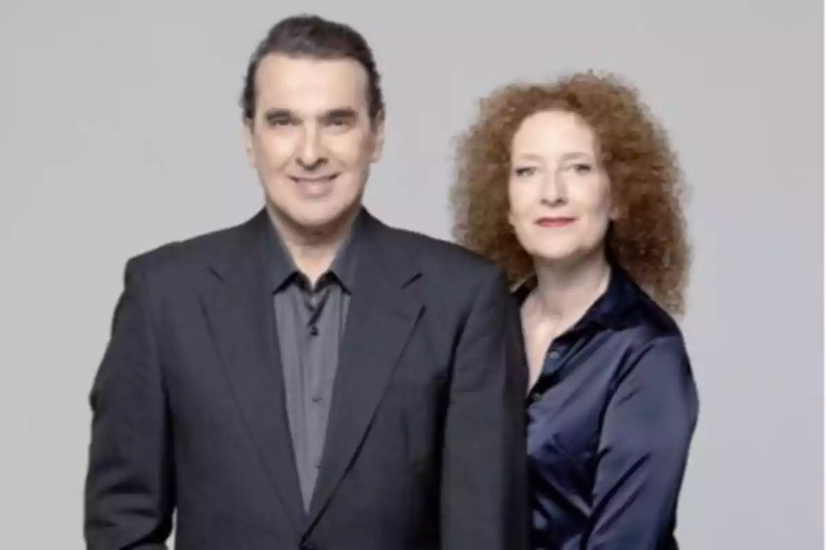 Dos personas posan juntas frente a un fondo gris, un hombre con saco oscuro y una mujer de cabello rizado y blusa azul oscuro. Dos personas posan juntas frente a un fondo gris, un hombre con saco oscuro y una mujer de cabello rizado y blusa azul oscuro.