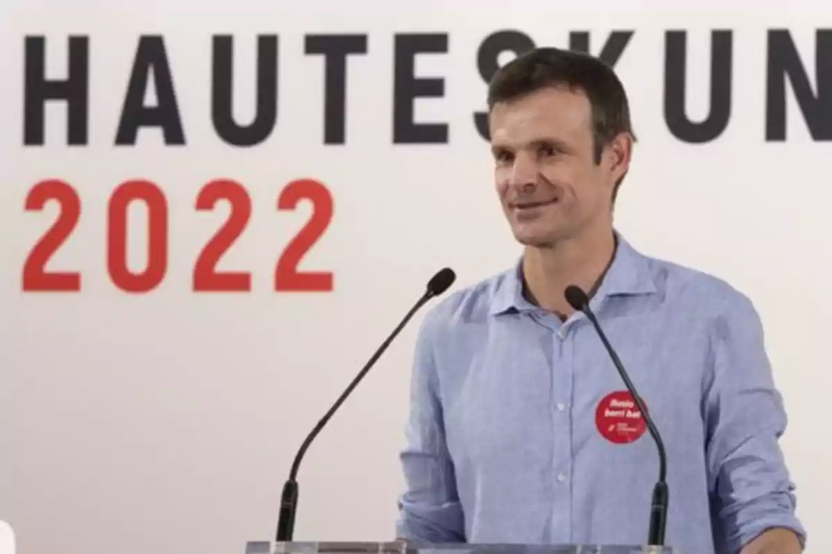 Hombre de camisa azul hablando en un podio con dos micrófonos y un fondo que muestra el texto HAUTESKUN 2022 Hombre de camisa azul hablando en un podio con dos micrófonos y un fondo que muestra el texto HAUTESKUN 2022