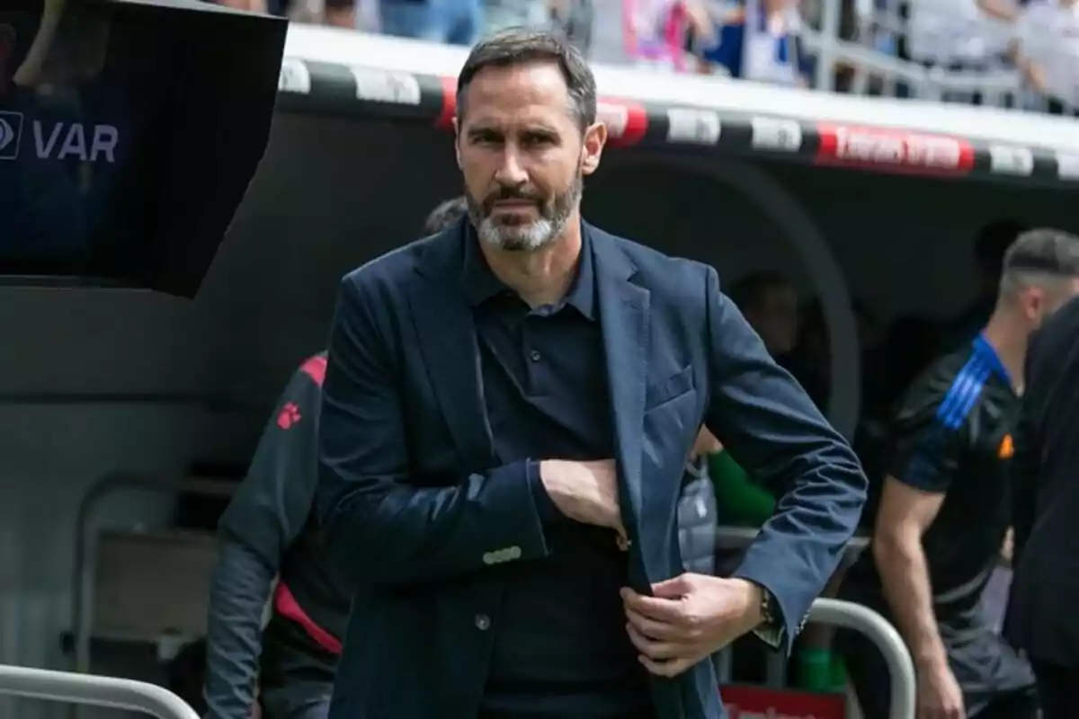 Hombre de barba y cabello corto con saco azul oscuro de pie en un estadio de fútbol junto a la zona de banquillo Hombre de barba y cabello corto con saco azul oscuro de pie en un estadio de fútbol junto a la zona de banquillo