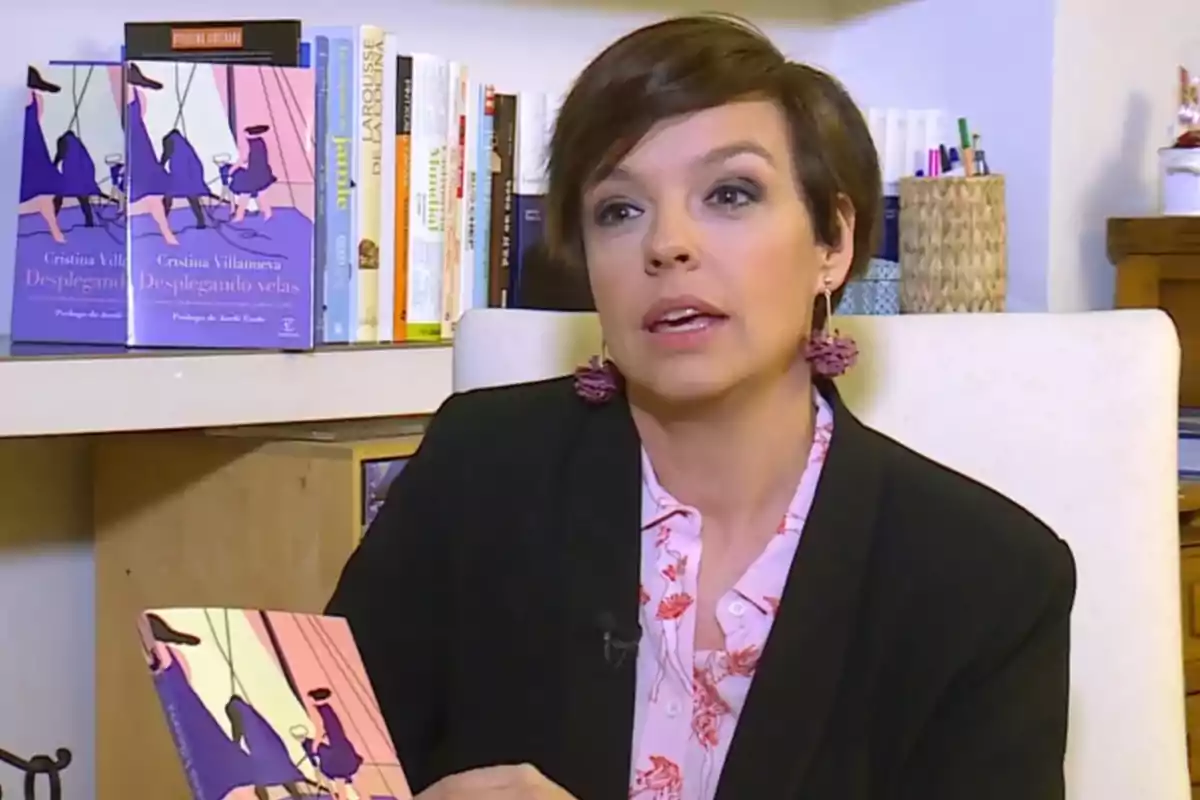 Mujer de cabello corto y oscuro sentada en una silla, sosteniendo un libro titulado Desplegando velas, con una estantería de libros al fondo Mujer de cabello corto y oscuro sentada en una silla, sosteniendo un libro titulado Desplegando velas, con una estantería de libros al fondo
