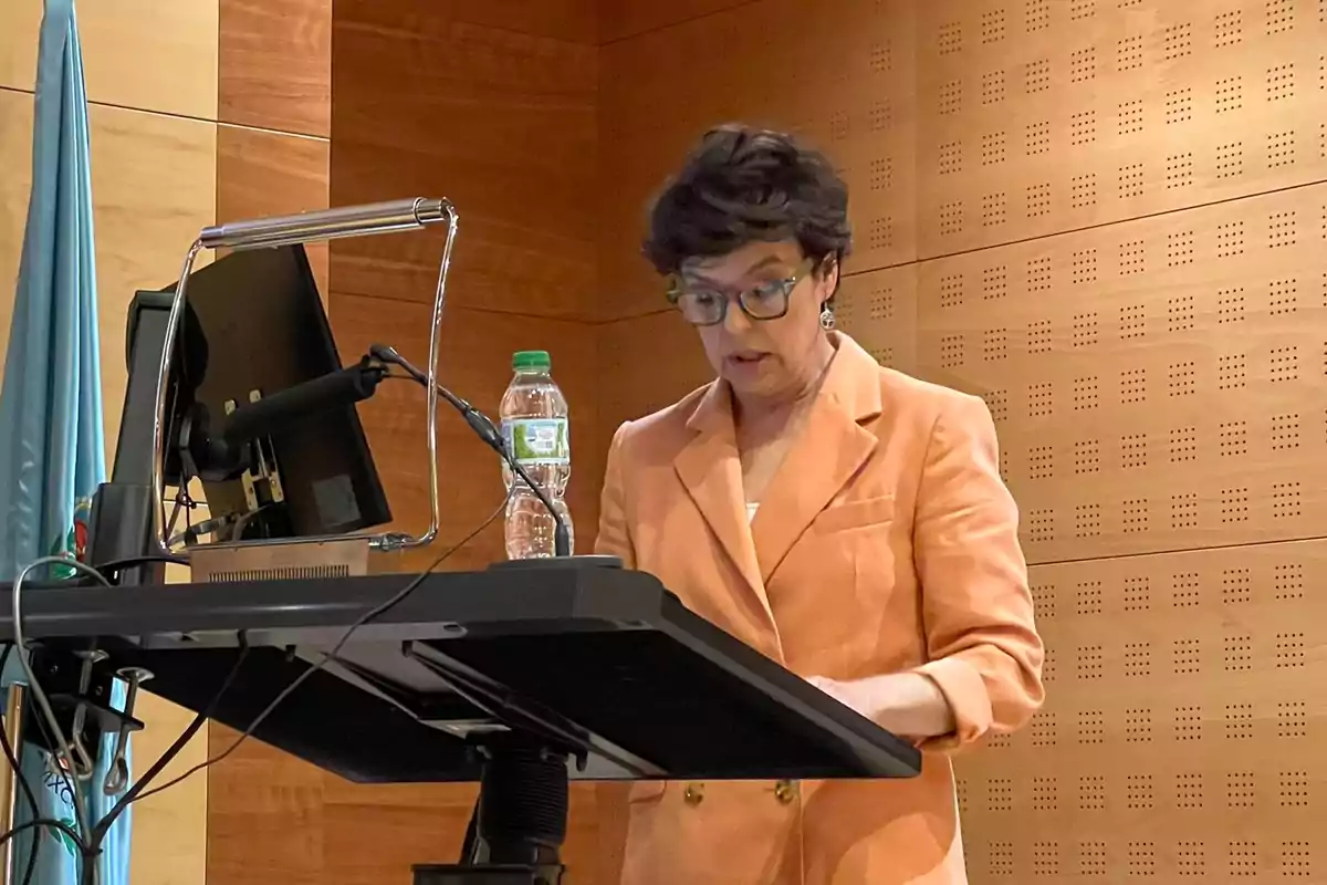 Mujer con gafas y chaqueta naranja hablando en un atril con una botella de agua y una pantalla en una sala de conferencias Mujer con gafas y chaqueta naranja hablando en un atril con una botella de agua y una pantalla en una sala de conferencias