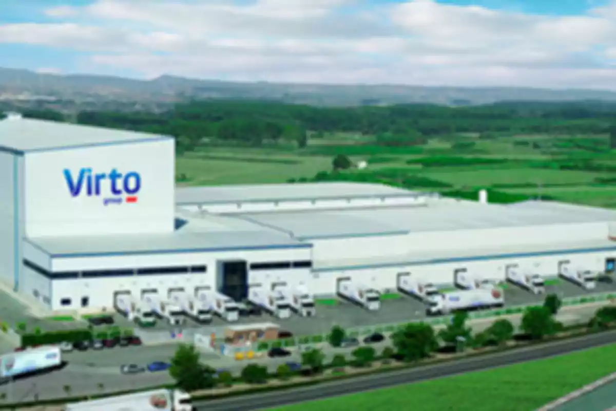 Vista aérea de una gran nave industrial blanca con el logo de Virto en la fachada principal rodeada de áreas verdes y varios camiones estacionados en los muelles de carga Vista aérea de una gran nave industrial blanca con el logo de Virto en la fachada principal rodeada de áreas verdes y varios camiones estacionados en los muelles de carga