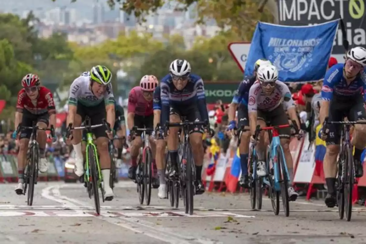 Ciclistas compitiendo en una carrera mientras cruzan la línea de meta con público y banderas al fondo Ciclistas compitiendo en una carrera mientras cruzan la línea de meta con público y banderas al fondo