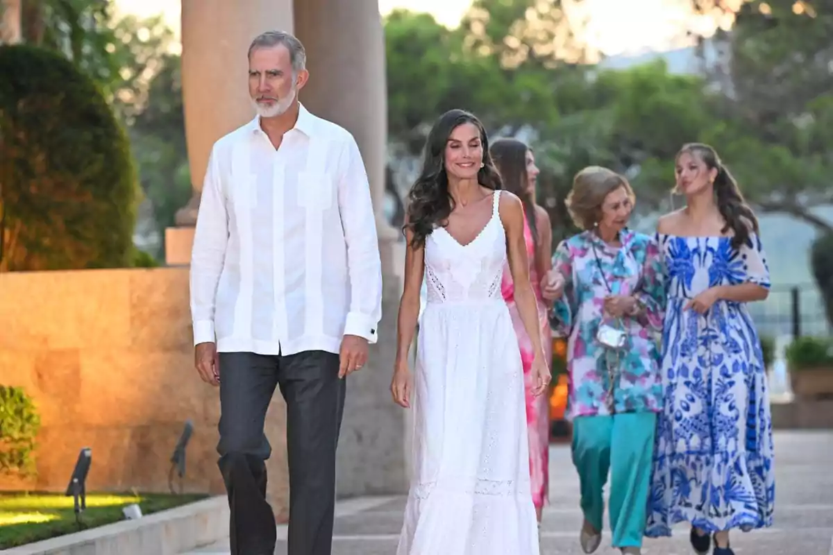 Un hombre y una mujer caminan juntos al frente mientras otras tres mujeres conversan detrás de ellos en un entorno al aire libre con árboles y columnas Un hombre y una mujer caminan juntos al frente mientras otras tres mujeres conversan detrás de ellos en un entorno al aire libre con árboles y columnas