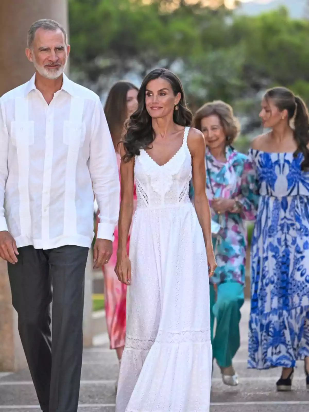 Un hombre y una mujer caminan juntos al aire libre, ambos vestidos elegantemente de blanco, seguidos por otras personas con ropa colorida Un hombre y una mujer caminan juntos al aire libre, ambos vestidos elegantemente de blanco, seguidos por otras personas con ropa colorida
