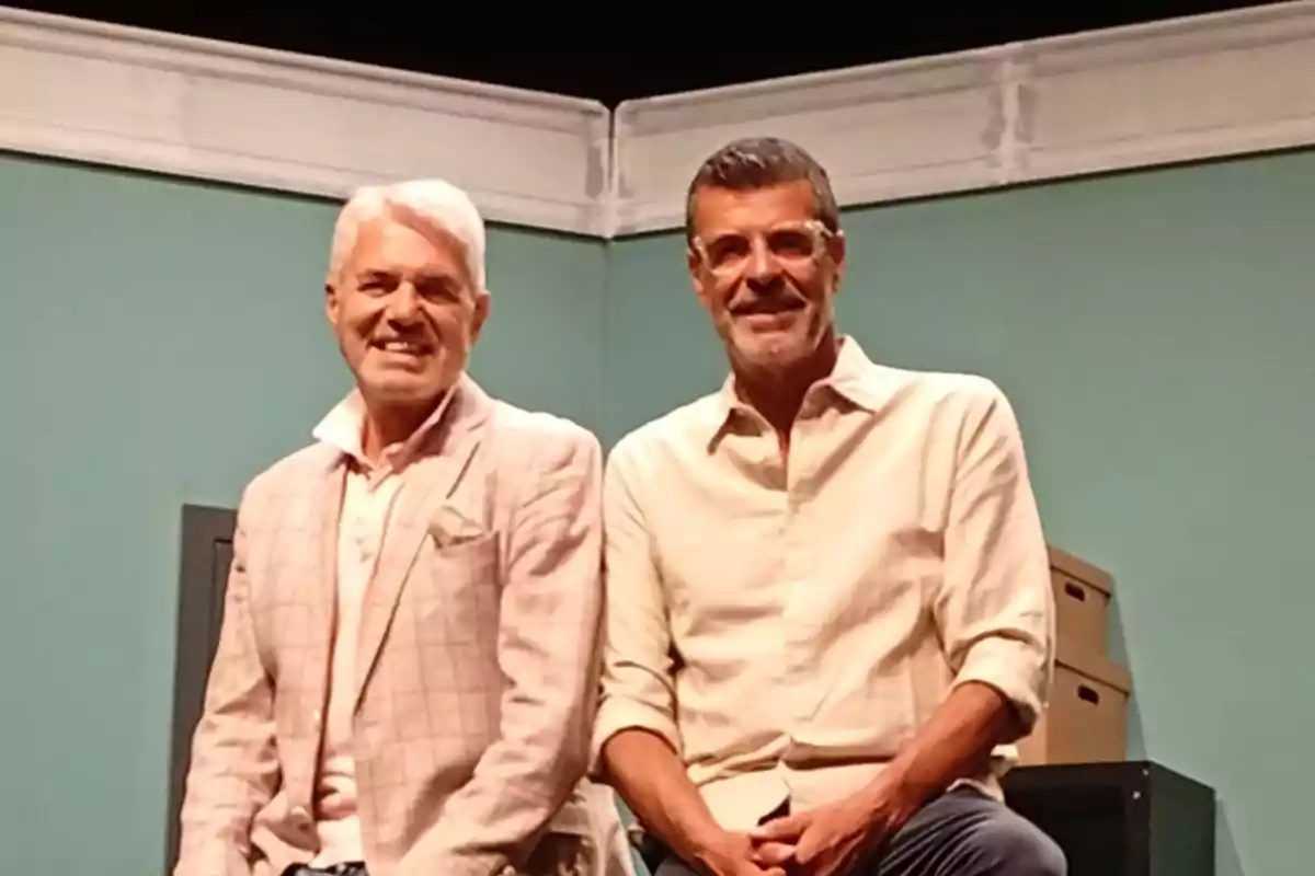 Dos hombres de cabello canoso y barba posan sonrientes en un escenario con paredes verdes y cajas de cartón al fondo