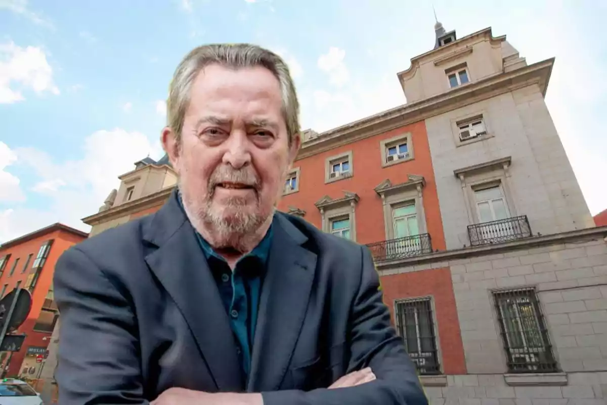 Hombre mayor de cabello canoso y barba con saco oscuro posando frente a un edificio antiguo de ladrillo rojo y piedra en una ciudad