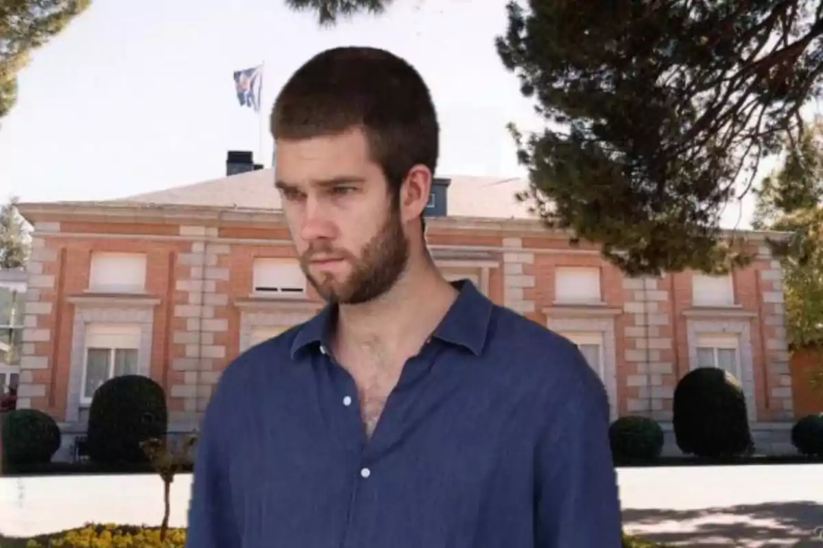 Un hombre de barba y cabello corto con camisa azul está de pie frente a una casa grande de ladrillo rodeada de árboles y arbustos.